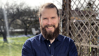 Henrik Johansson, man med mörkt hår och skägg, blå skjorta med soli trädgård i bakgrunden, ler mot kameran.